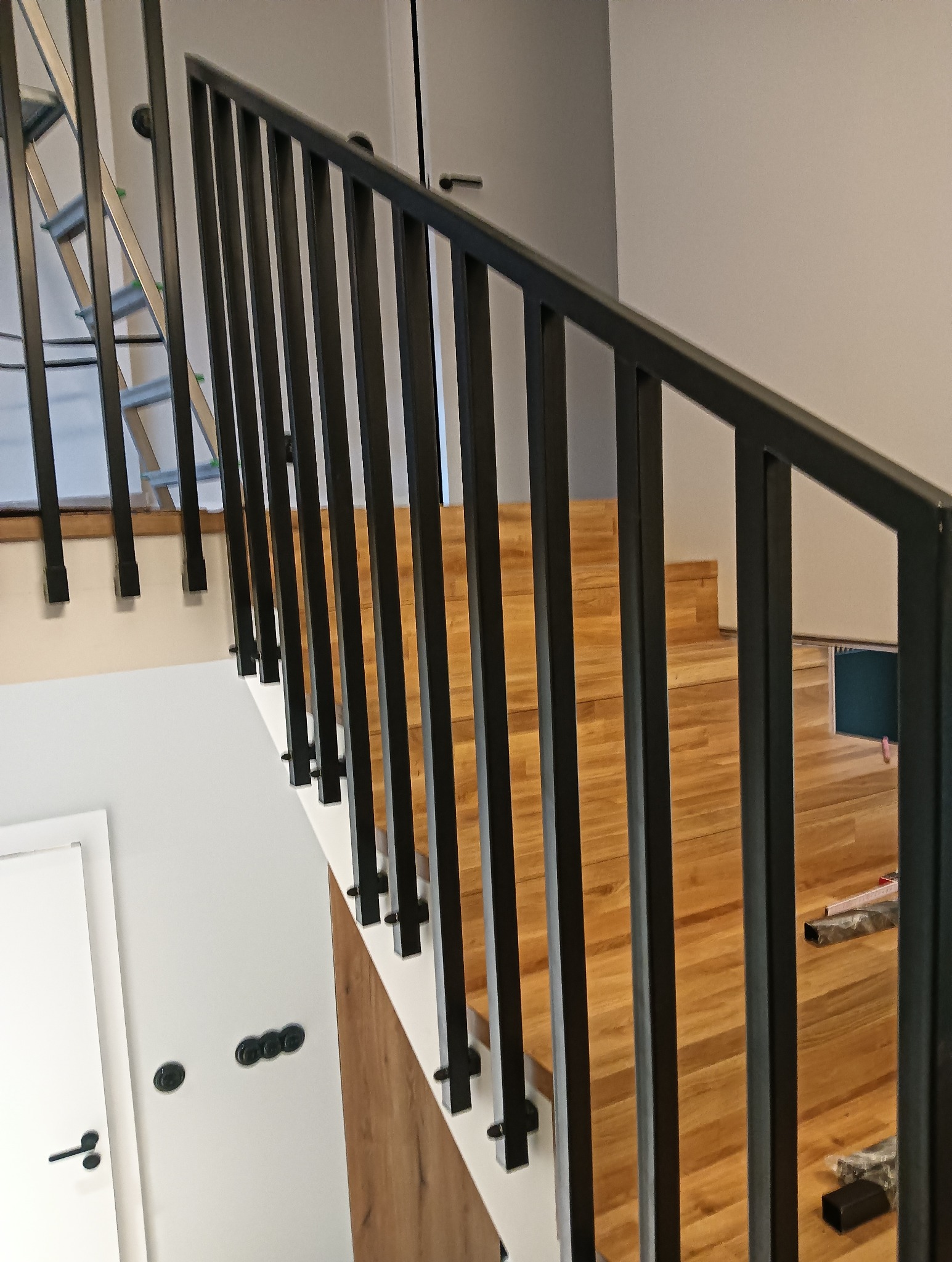 Nowoczesna, czarna poręcz schodowa z pionowymi szczeblami na drewnianych schodach. Widoczny fragment drabiny w tle. Minimalistyczny design.