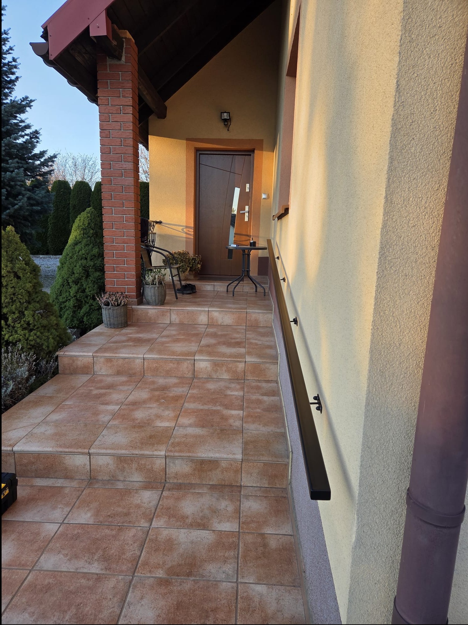 Zewnętrzne schody wejściowe z brązowymi płytkami i czarną poręczą zamontowaną na ścianie budynku. Widoczne rośliny w donicach i fragment elewacji w ciepłym odcieniu.