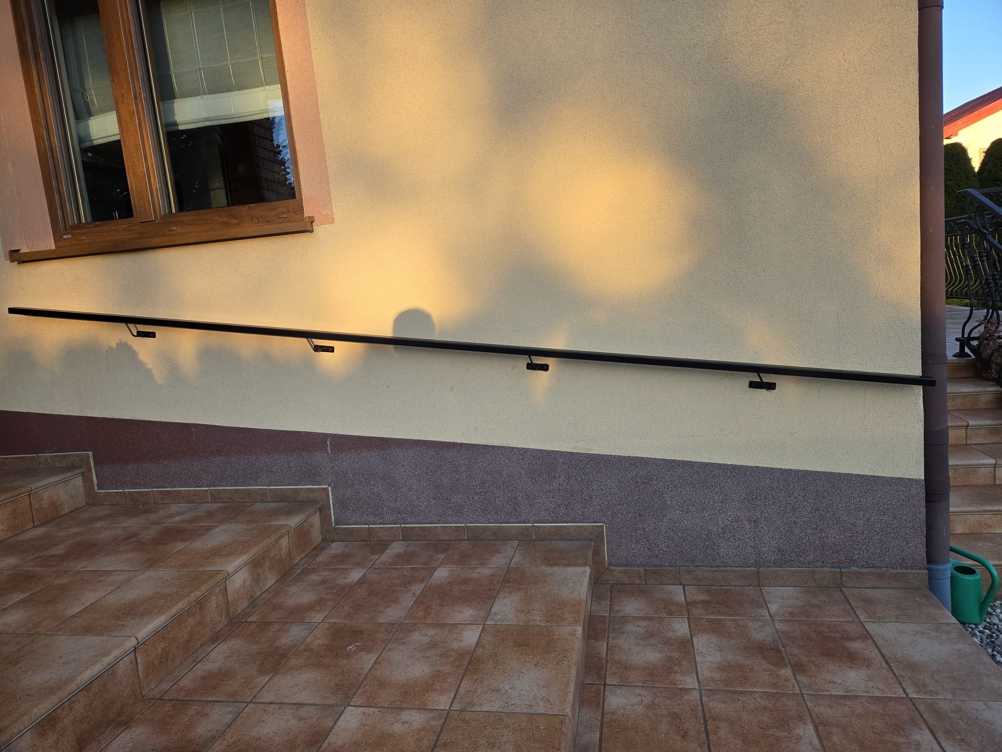 Czarna, metalowa poręcz zamontowana na ścianie budynku obok schodów zewnętrznych z brązowych płytek. Widoczne okno i fragment balustrady schodów.