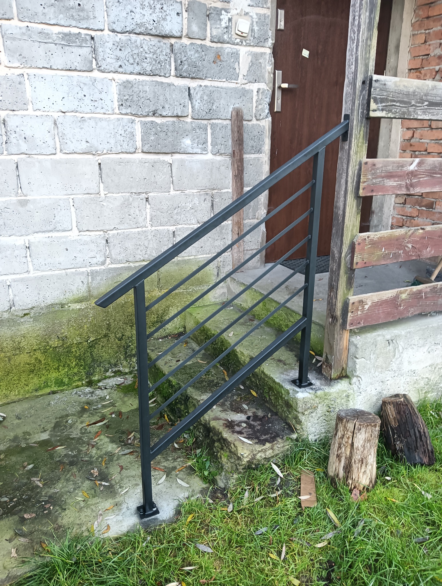 Nowoczesna, czarna, metalowa konstrukcja schodowa z poziomymi prętami, zamontowana przy wejściu do budynku z szarej cegły. Widoczne elementy drewniane i trawa.