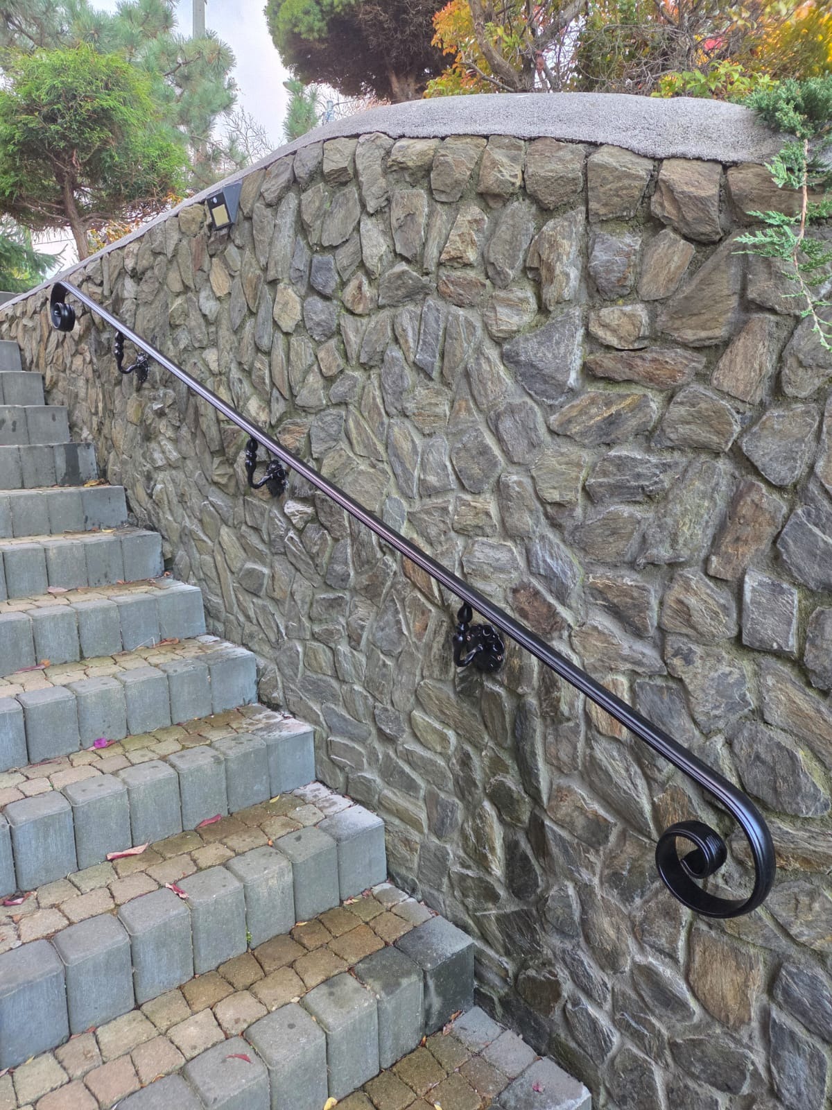 Czarna, metalowa poręcz schodowa z ozdobnym zakończeniem, zamontowana na kamiennej ścianie wzdłuż stopni z kostki brukowej. Widok z dołu, oświetlenie naturalne.