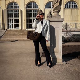 Alicja Kamińska PS Stylistka - Kobieta w stylizacji z białym żakietem, czarnymi spodniami z rozcięciami i brązową torebką, pozująca obok rzeźby na tle budynku z kolumnami.