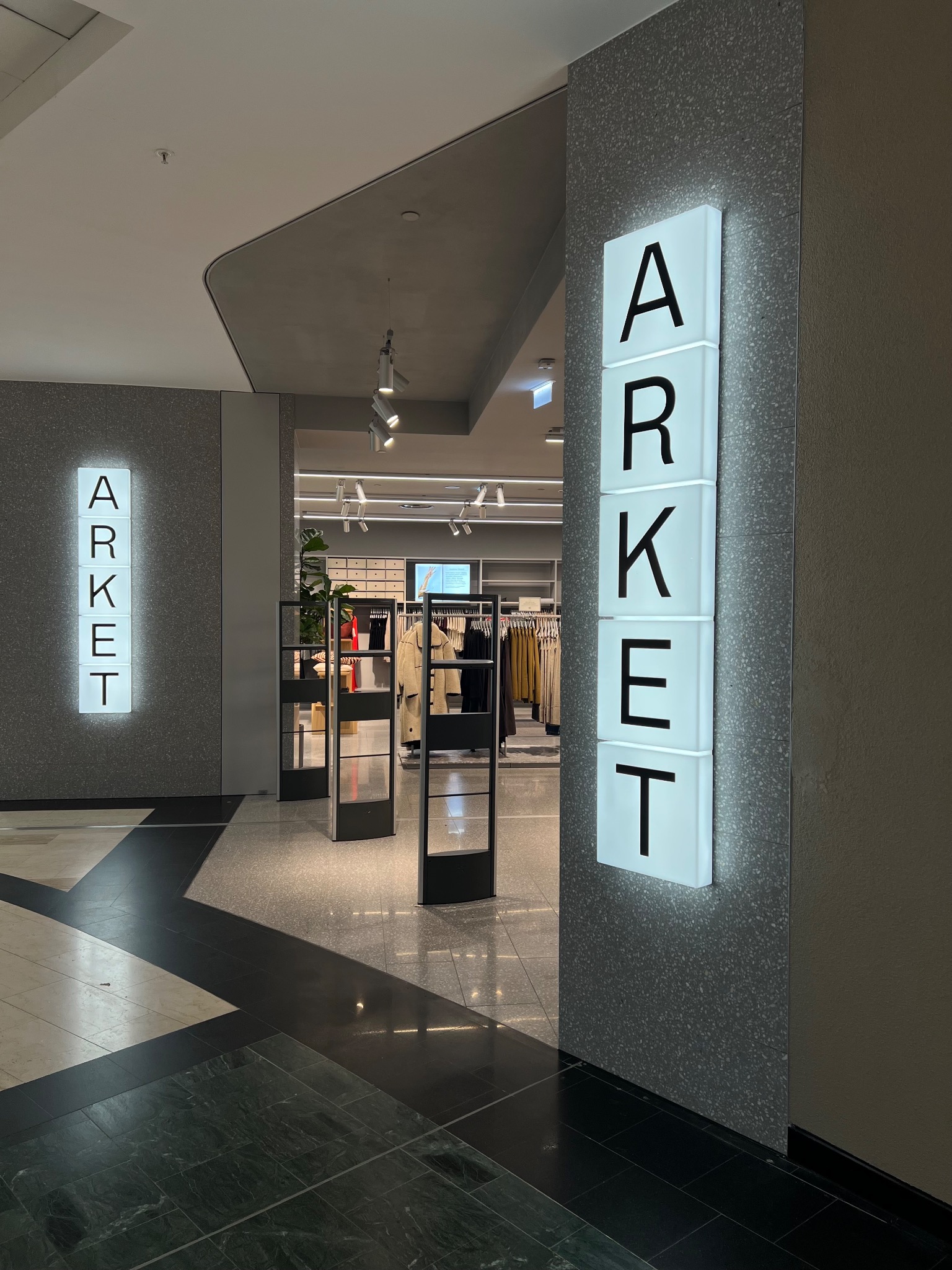 Wejście do sklepu odzieżowego Arket z podświetlanym logo na ścianie i widocznymi w głębi wieszakami z ubraniami.