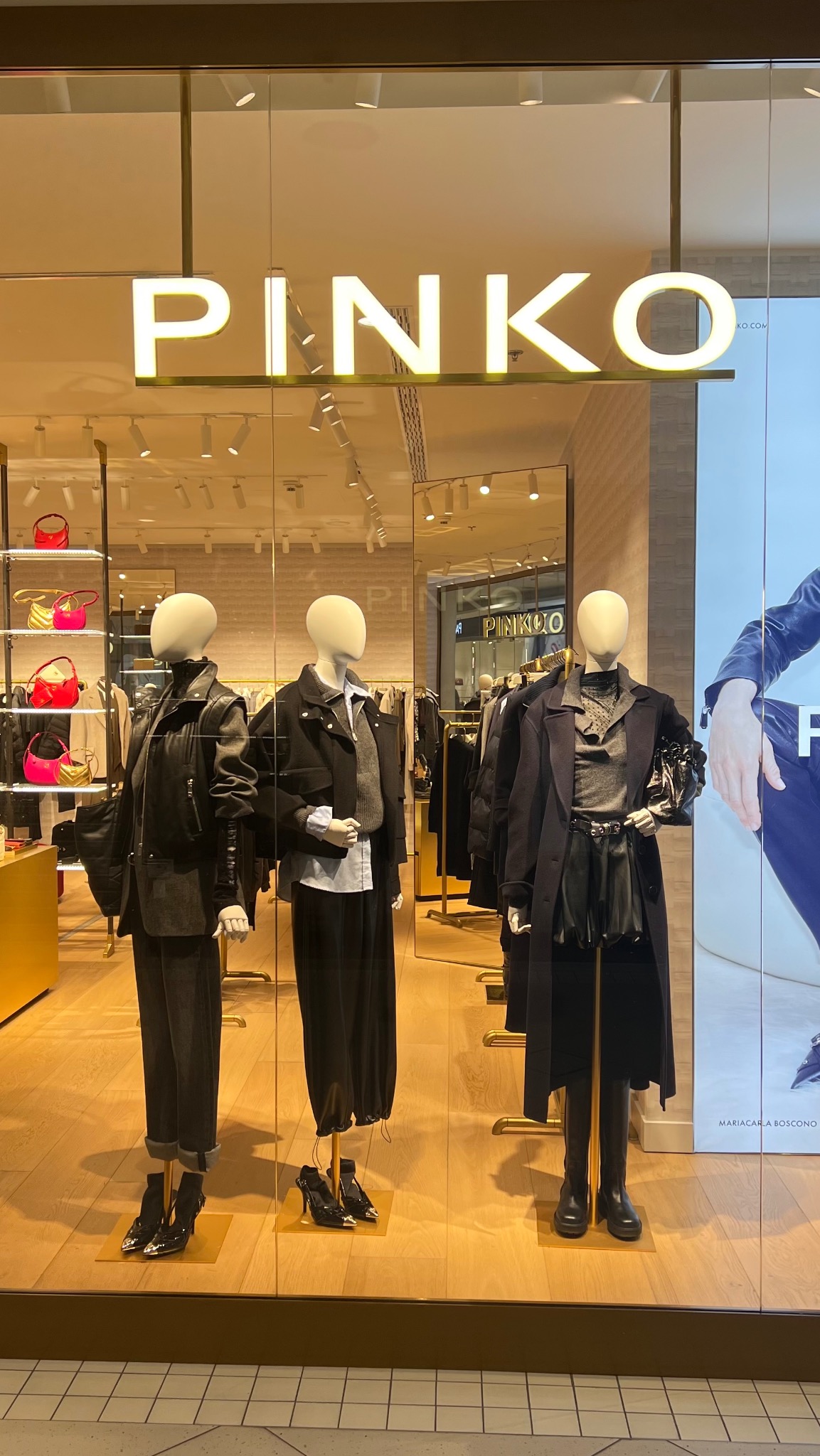 Witryna sklepu Pinko z trzema manekinami prezentującymi stylizacje w odcieniach czerni i szarości, podświetlony neonowy napis marki, w tle widoczne półki z torebkami.