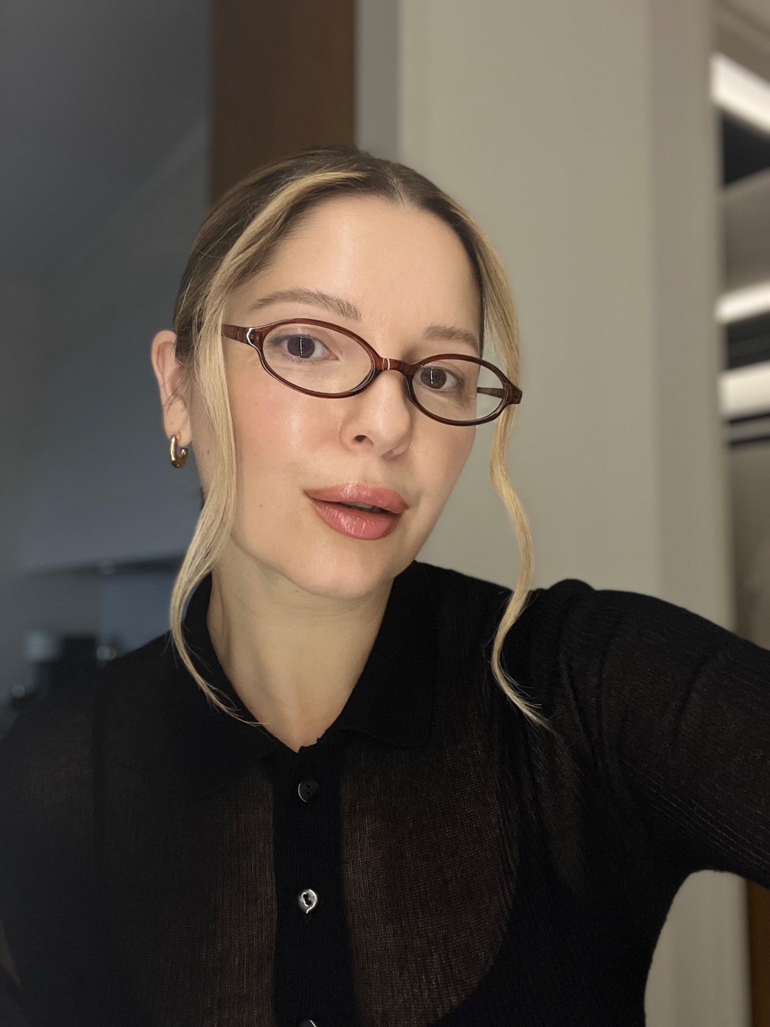 Portret kobiety w okularach o owalnych oprawkach, z blond pasemkami i makijażem nude, ubrana w czarną bluzkę z guzikami.