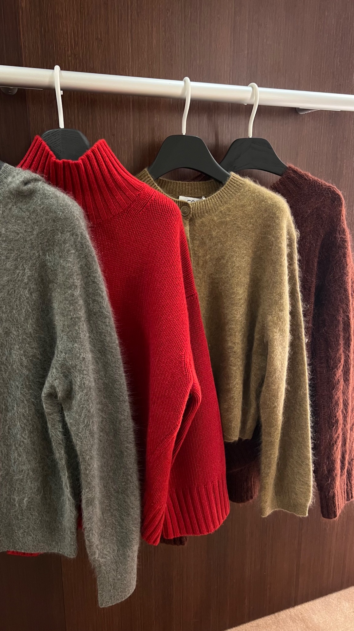 Rząd kolorowych swetrów z długim włosiem, wiszących na czarnych wieszakach na tle drewnianej ściany.
