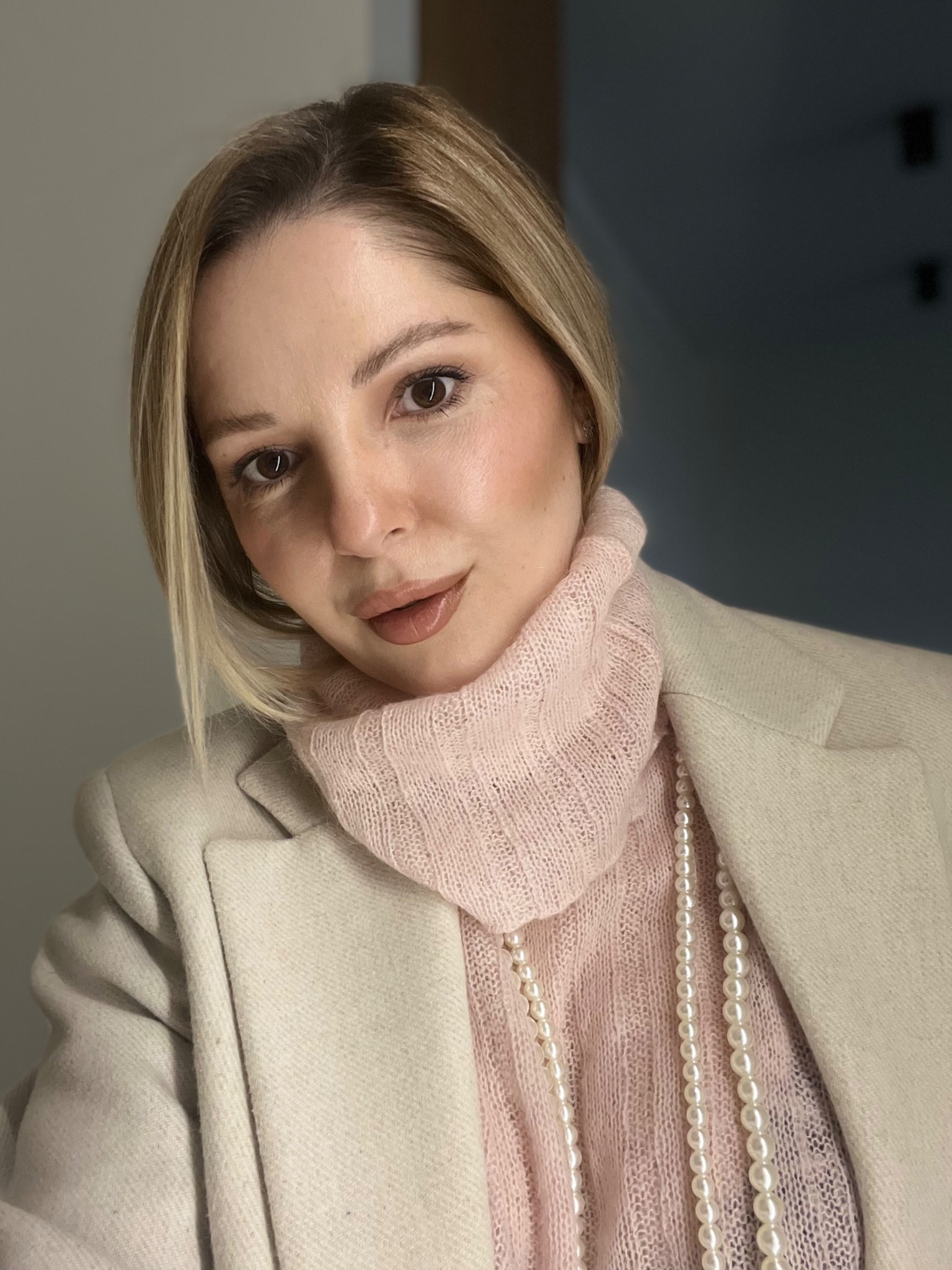 Portret kobiety z blond włosami w różowym swetrze z golfem i kremowym płaszczu, z perłami, delikatny makijaż.