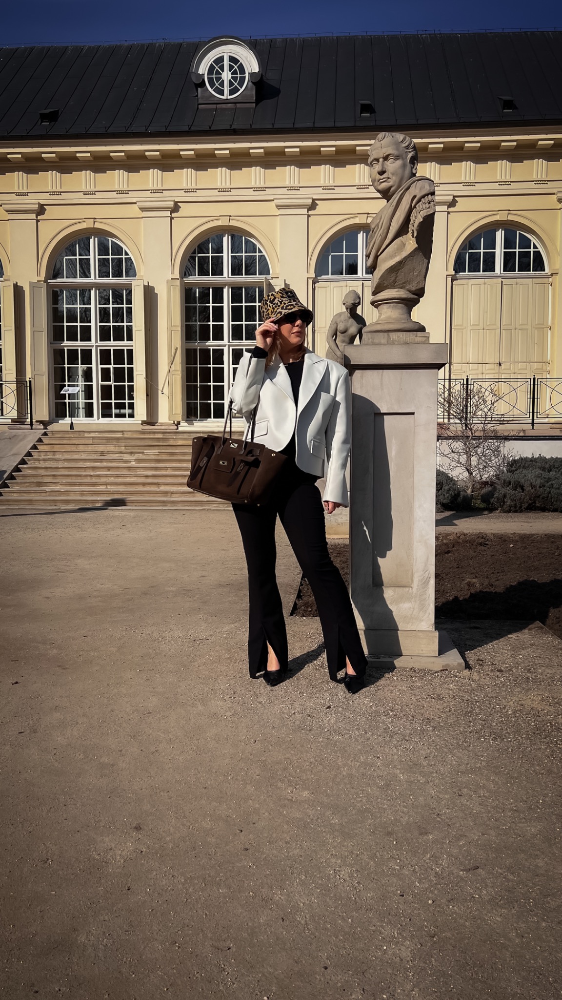 Kobieta w stylizacji z białym żakietem, czarnymi spodniami z rozcięciami i brązową torebką, pozująca obok rzeźby na tle budynku z kolumnami.