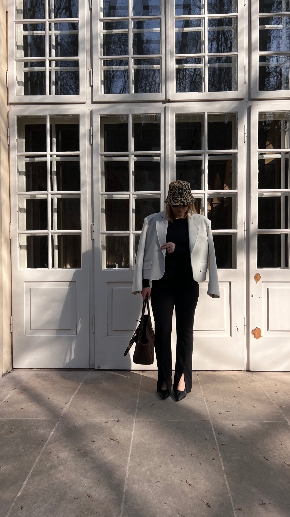 Kobieta w modnej stylizacji w Warszawie: czarne spodnie z rozcięciem, czarny top, jasna marynarka i kapelusz w panterkę, stojąca na tle białych drzwi z licznymi szybami.