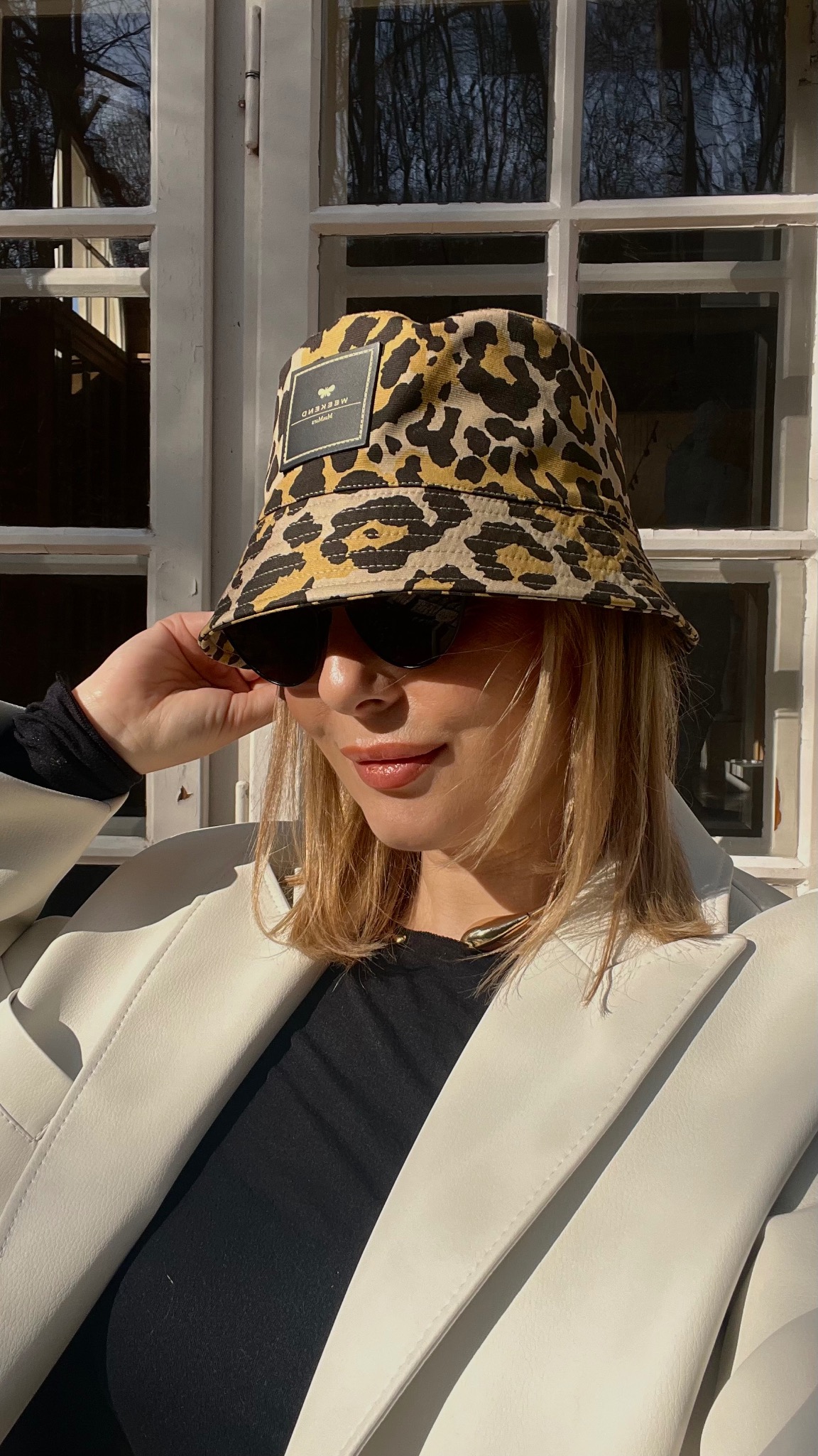 Kobieta w okularach przeciwsłonecznych i kapeluszu typu bucket hat w panterkę, ubrana w białą marynarkę, pozuje na tle okna.