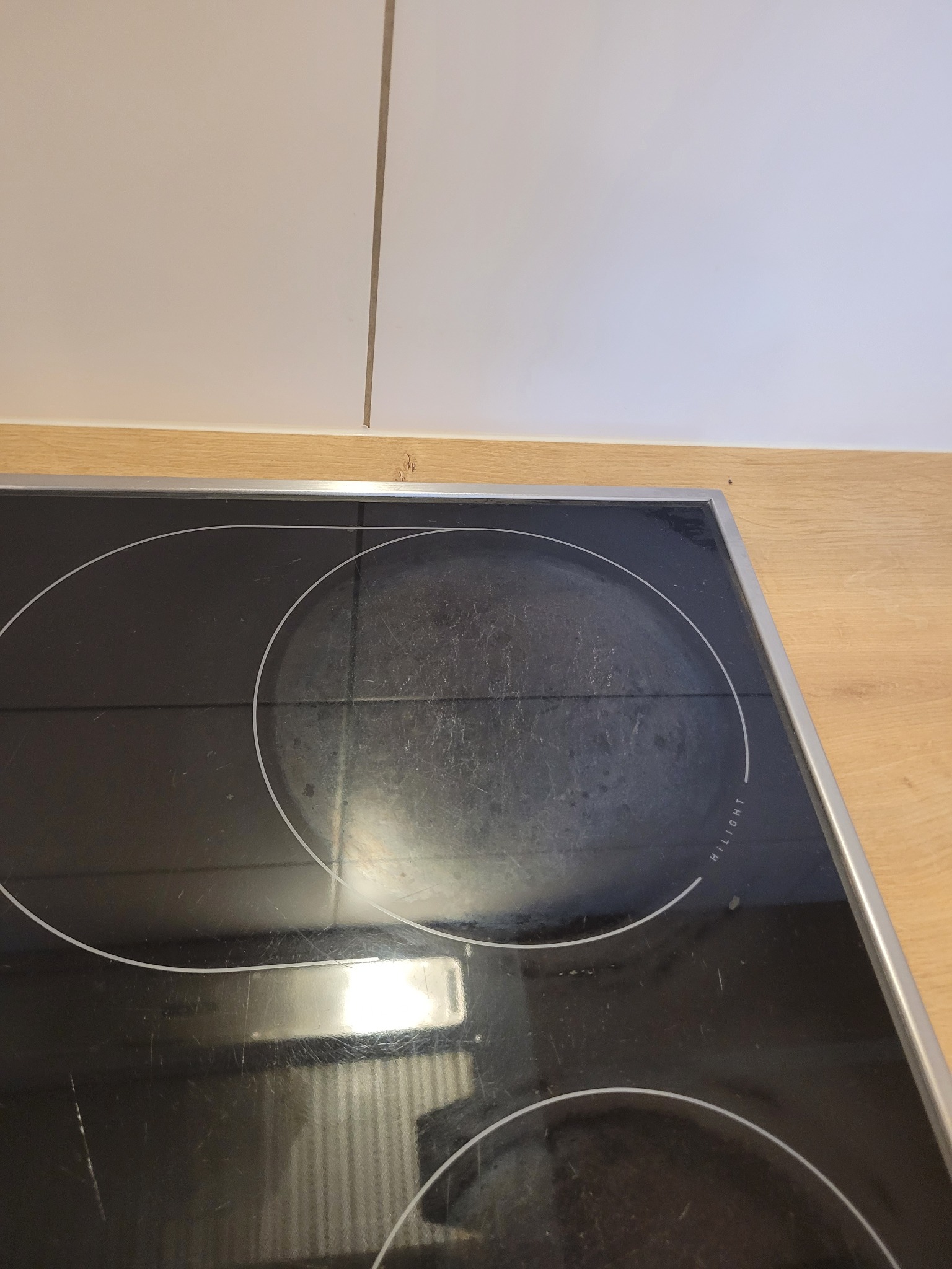 Brudna, czarna płyta indukcyjna z widocznymi śladami użytkowania i zaschniętymi plamami, umieszczona w blacie kuchennym o jasnym, drewnopodobnym wykończeniu, z białymi szafkami w tle.