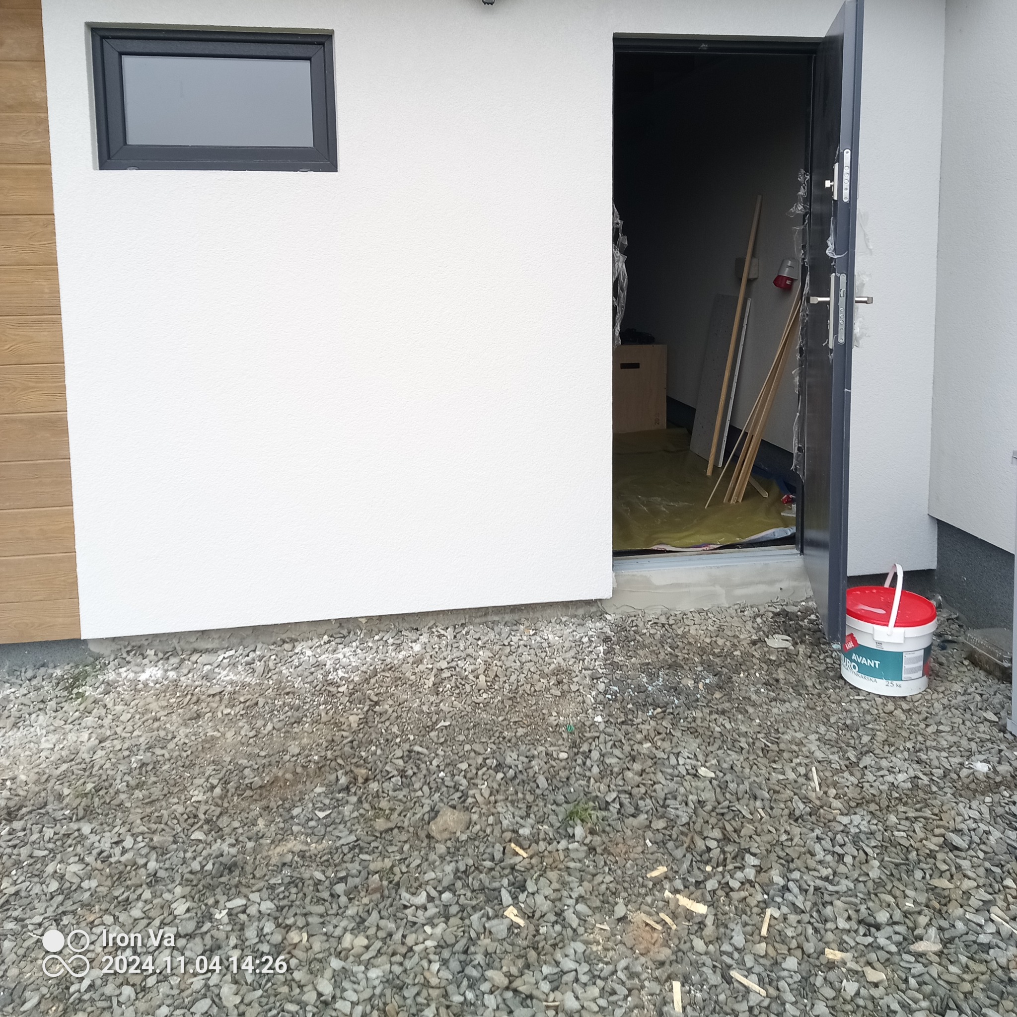 Biała fasada budynku z oknem i otwartymi drzwiami, widoczny kubeł z farbą na żwirowym podłożu, zabezpieczenie folią malarską wewnątrz.