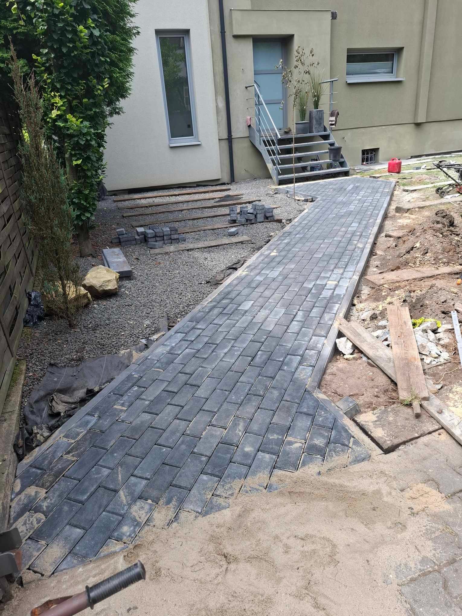Układanie szarej kostki brukowej na ścieżce prowadzącej do domu, widoczne narzędzia i piasek, prace w toku.