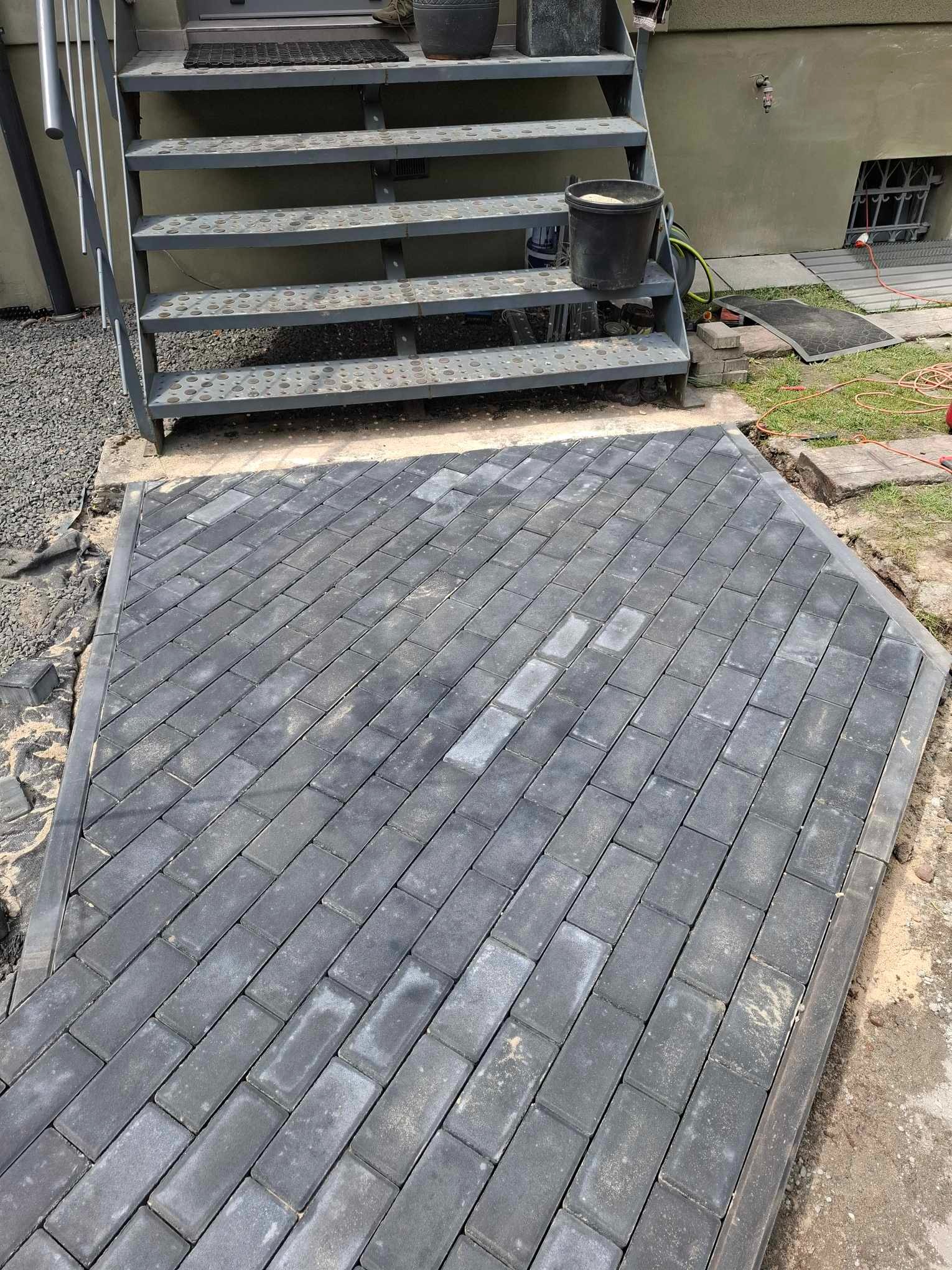Ułożona kostka brukowa w odcieniach szarości, ułożona pod kątem przed metalowymi schodami wejściowymi do budynku, widoczne niedociągnięcia w wykończeniu i pozostałości piasku.