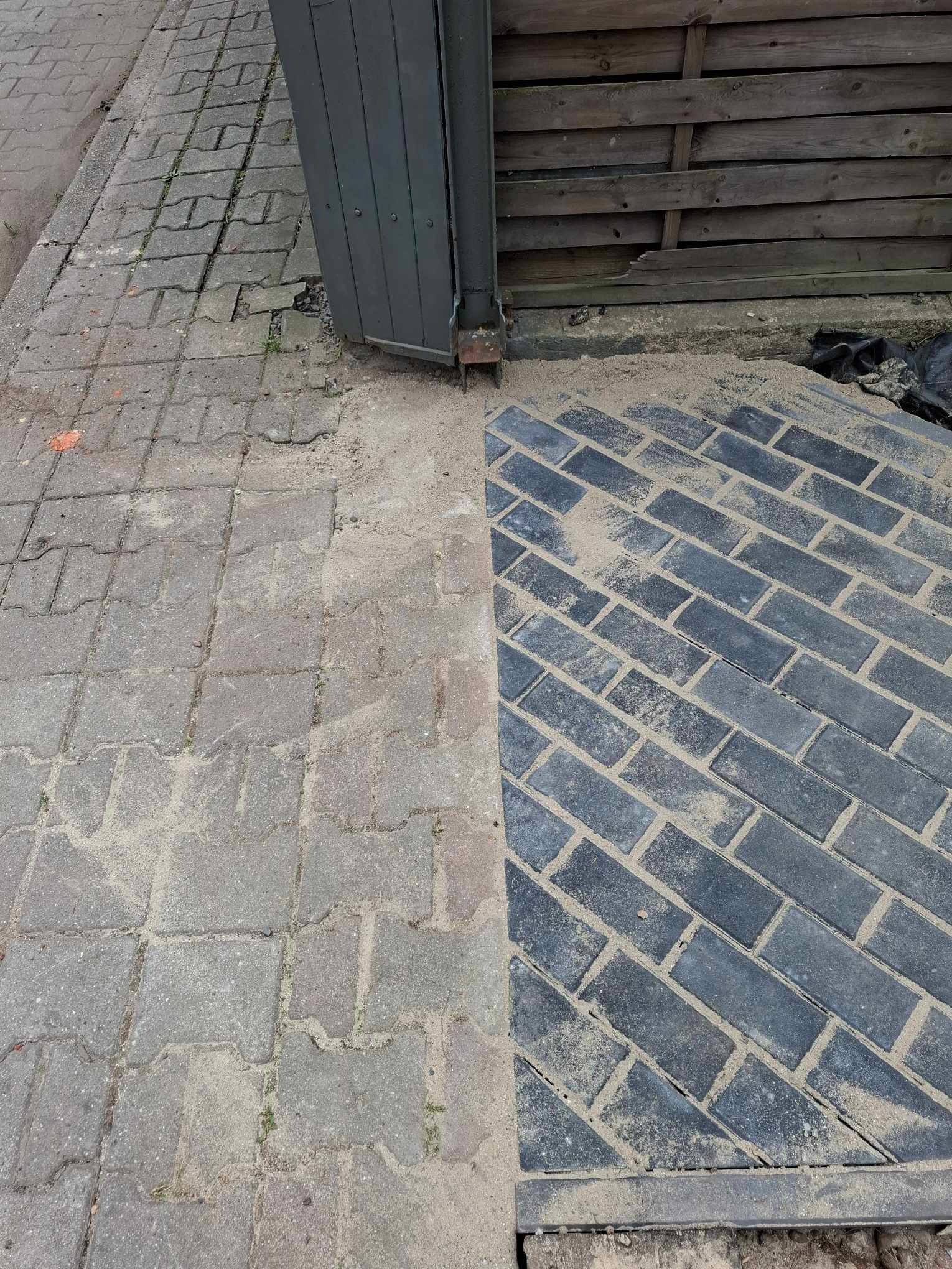 Połowa chodnika z szarej kostki brukowej, druga połowa z nowo ułożonej, ciemnej kostki, zasypanej piaskiem, przy drewnianym płocie.
