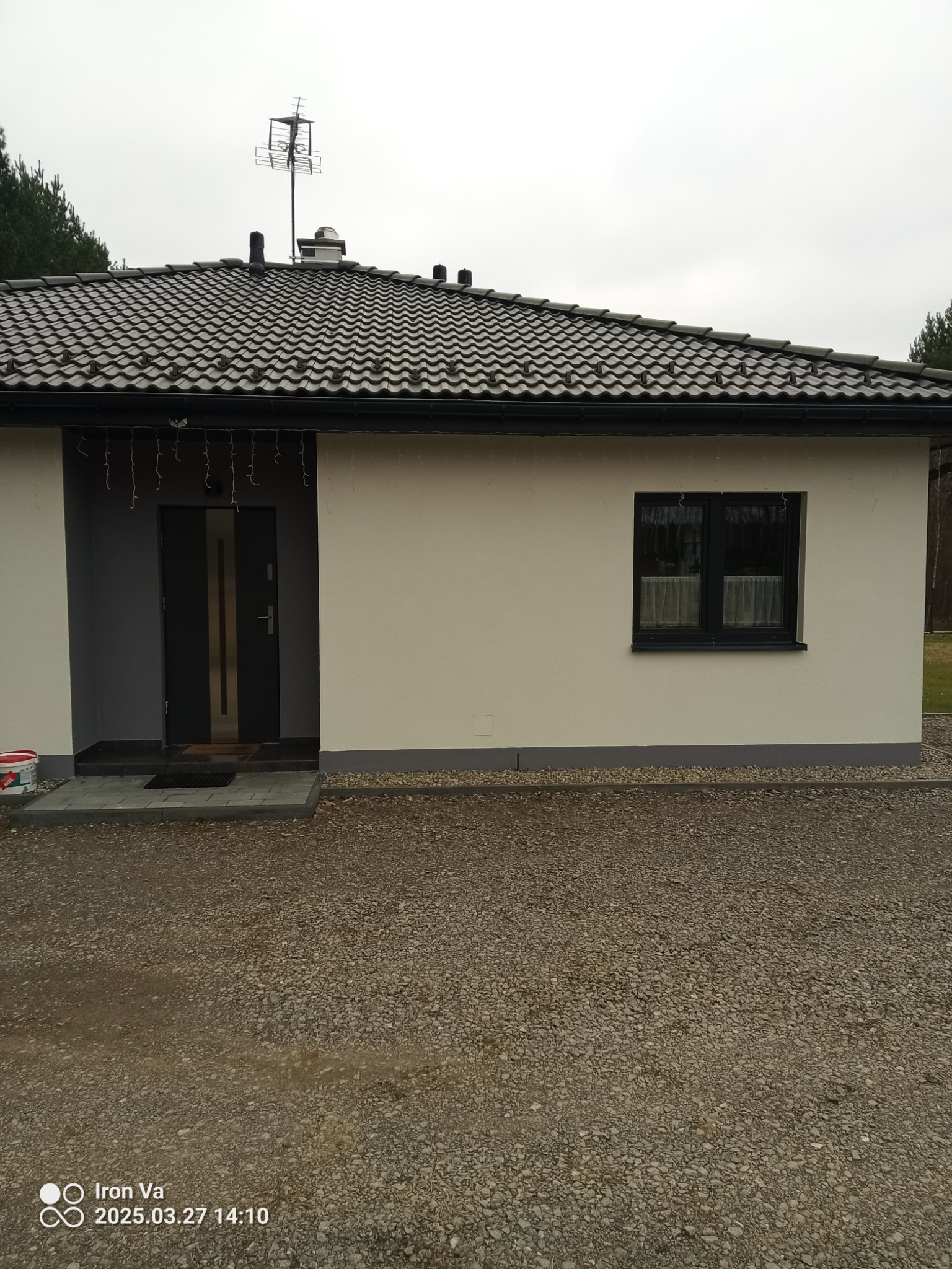 Świeżo otynkowany, parterowy dom jednorodzinny z szarą stolarką okienną i drzwiową, widok z zewnątrz na fasadę i podjazd wysypany żwirem.