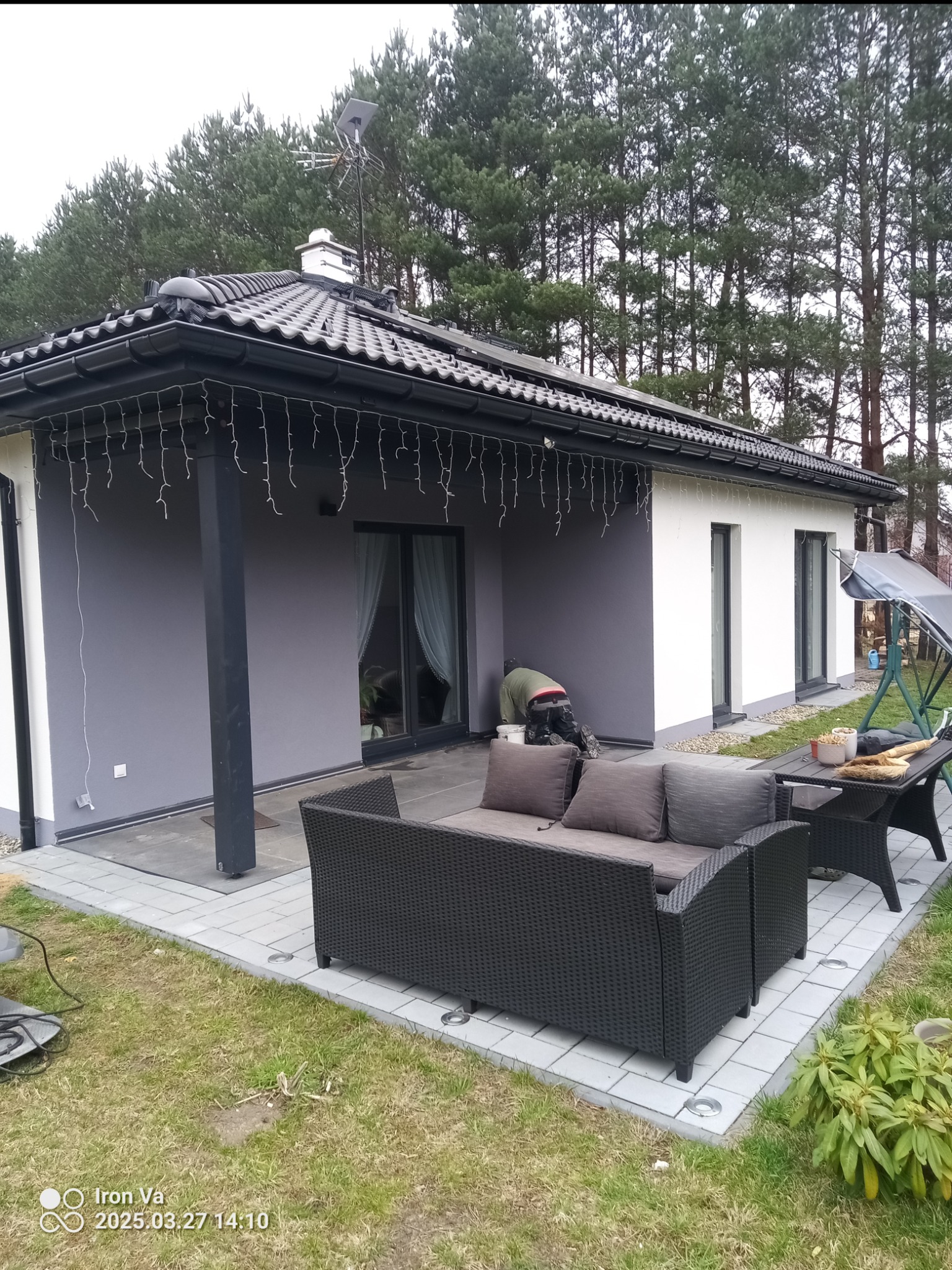 Pracownik w trakcie malowania elewacji domu jednorodzinnego na szaro, widoczny fragment tarasu z meblami ogrodowymi i narzędzia malarskie obok.