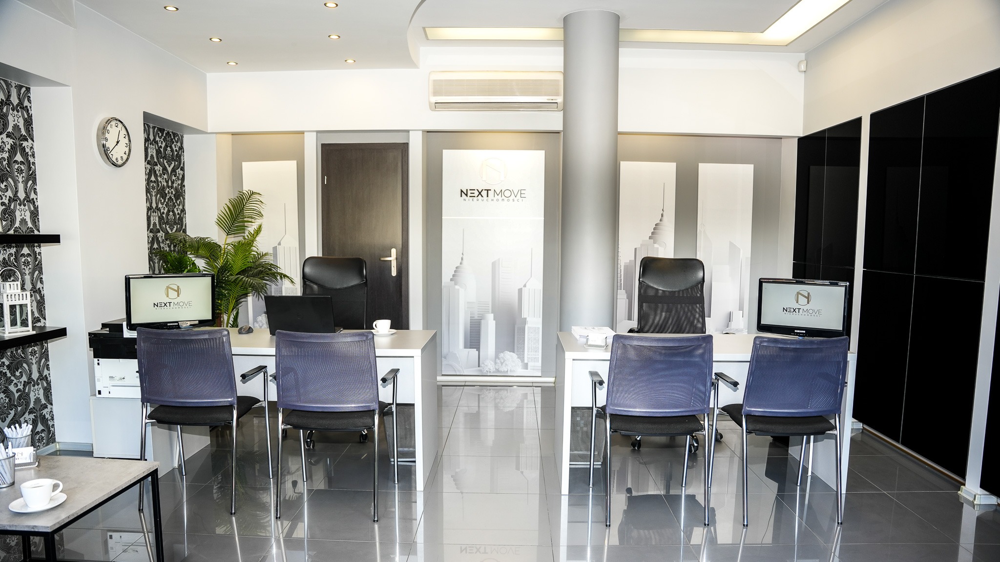 Nowoczesne biuro agencji nieruchomości z dwoma stanowiskami pracy, monitorami z logo firmy, rośliną doniczkową, zegarem na ścianie i błyszczącą podłogą.