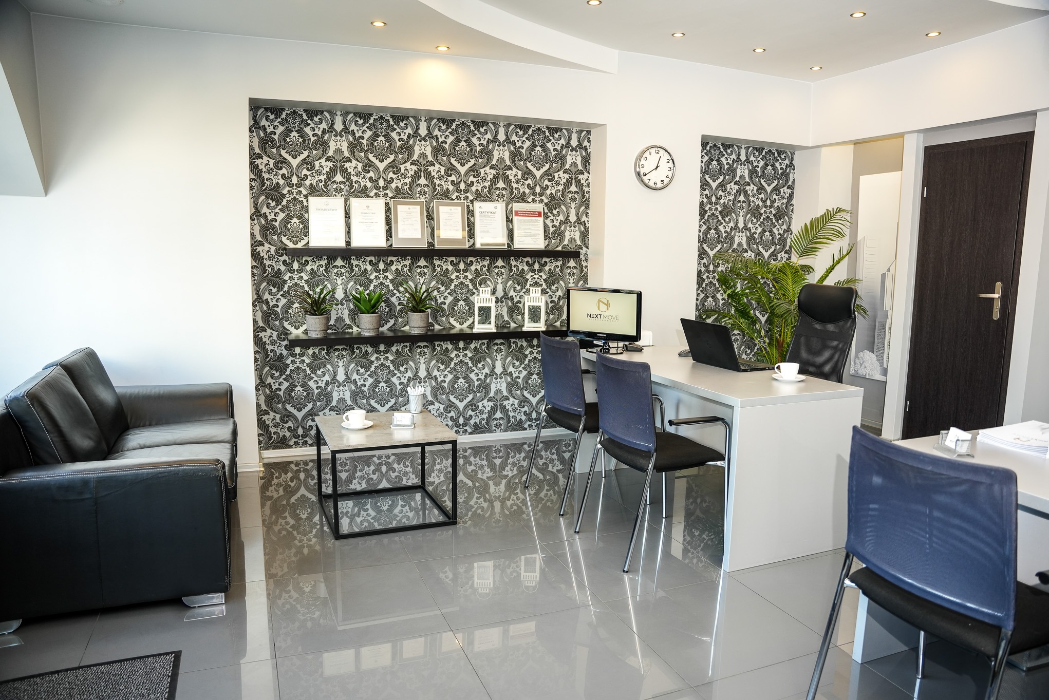 Nowoczesne biuro z czarną skórzaną sofą, stolikiem kawowym z filiżankami, biurkiem z laptopem i monitorem z logo firmy, ozdobną tapetą i certyfikatami na ścianie.