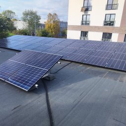Avotech - Instalacja fotowoltaiczna na płaskim dachu budynku mieszkalnego, widoczne kable i elementy montażowe, w tle budynki i drzewa.