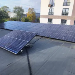 Instalacja fotowoltaiczna na płaskim dachu budynku mieszkalnego, widoczne kable i elementy montażowe, w tle budynki i drzewa.