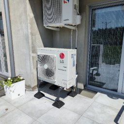 Avotech - Zewnętrzna jednostka pompy ciepła LG Therma V na metalowym stelażu, zainstalowana na tarasie obok okna balkonowego, z widoczną jednostką wewnętrzną na ścianie. W tle donice z kwiatami.