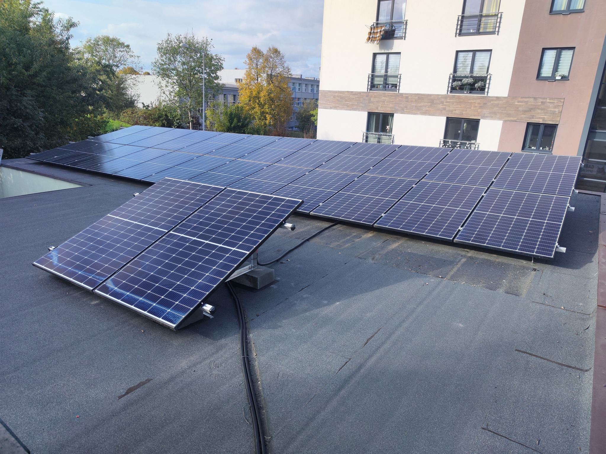 Instalacja fotowoltaiczna na płaskim dachu budynku mieszkalnego, widoczne kable i elementy montażowe, w tle budynki i drzewa.