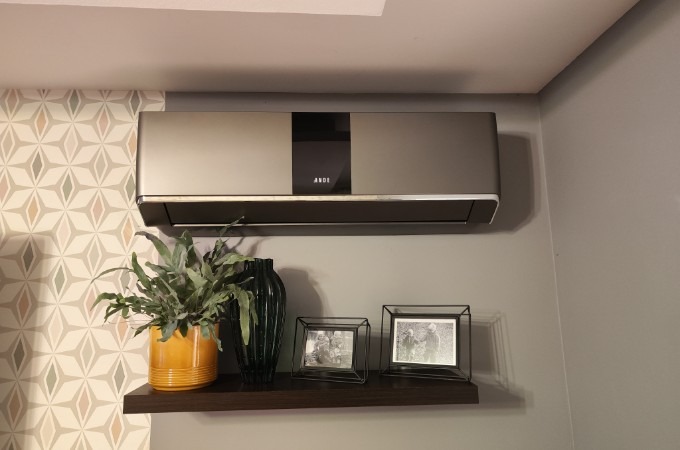 Szary klimatyzator ścienny marki ANDE, zamontowany nad półką z dekoracjami: żółtą doniczką z paprotką, ciemnozielonym wazonem i dwoma metalowymi ramkami ze zdjęciami.