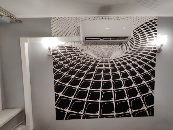 Biała jednostka klimatyzacyjna zamontowana na szarej ścianie z czarno-białą tapetą z iluzją optyczną tunelu, oświetlona kinkietami.