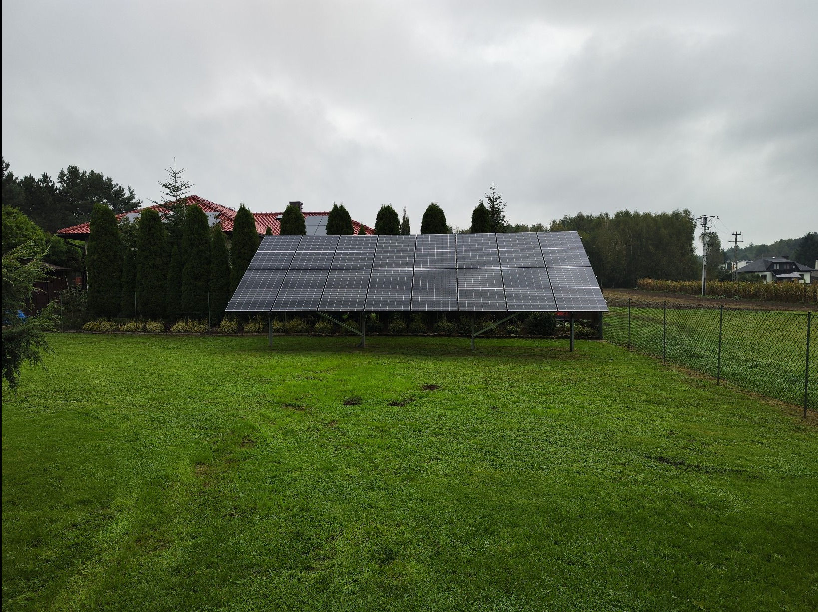 Wolnostojąca instalacja fotowoltaiczna na metalowej konstrukcji, umieszczona na trawniku przed domem z czerwoną dachówką w pochmurny dzień.