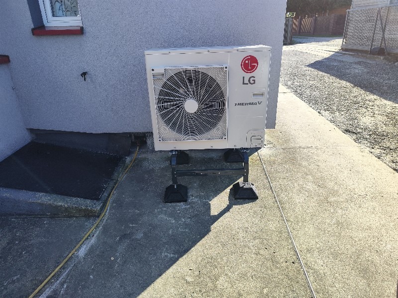 Biała pompa ciepła LG Therma V zainstalowana na czarnym, metalowym stelażu na zewnątrz budynku. Widoczny fragment elewacji i chodnika.