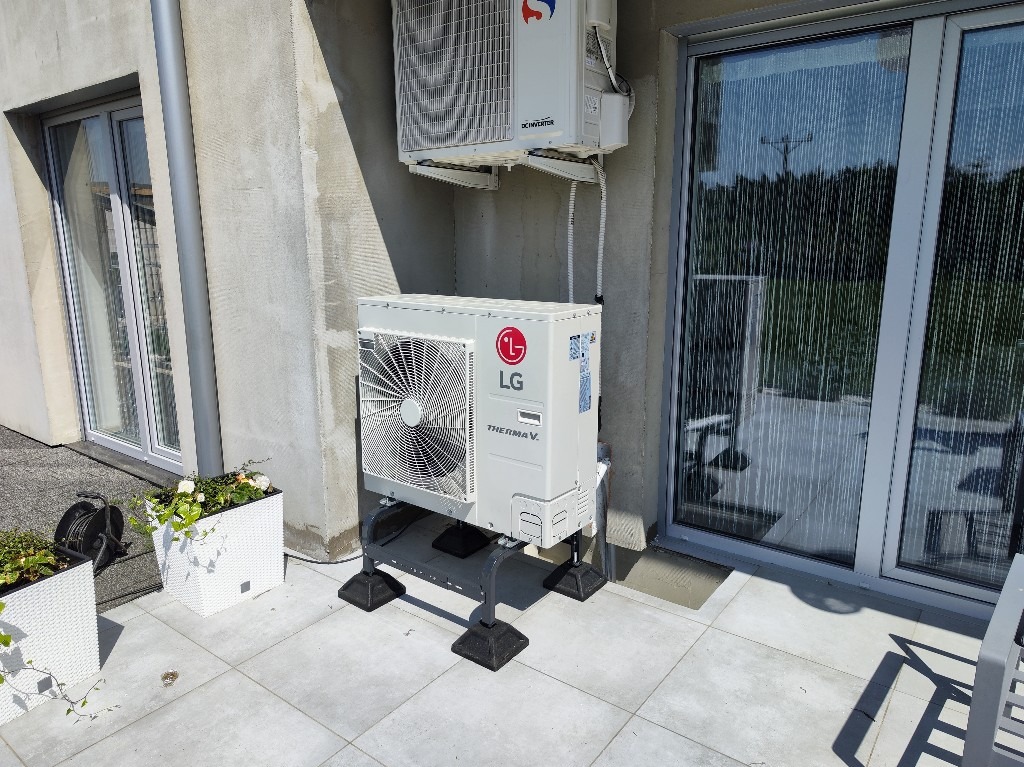 Zewnętrzna jednostka pompy ciepła LG Therma V na metalowym stelażu, zainstalowana na tarasie obok okna balkonowego, z widoczną jednostką wewnętrzną na ścianie. W tle donice z kwiatami.