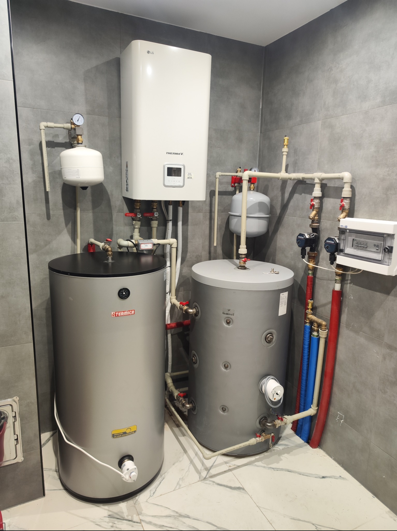 Kompletna instalacja grzewcza z dwoma zbiornikami Termica, pompą ciepła LG Therma V i osprzętem hydraulicznym na tle szarych płytek w łazience.