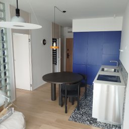 Serra's team Zakład Budowlany Rafał Serafin - Nowoczesne wnętrze mieszkania: okrągły stół z krzesłami, zabudowa kuchenna w kolorze niebieskim, parkiet i dekoracyjna ściana z luksferów. Widoczne lampy wiszące.