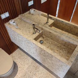 Nowoczesna łazienka z beżową mozaiką: umywalka nablatowa z dwoma kranami, lustro, toaleta. Drewniane panele na ścianie dodają ciepła. Widok z góry.