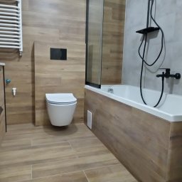 Nowoczesna łazienka z drewnopodobnymi płytkami, białą armaturą i czarną baterią prysznicową. Kabina prysznicowa z czarnymi profilami. Minimalistyczny design.