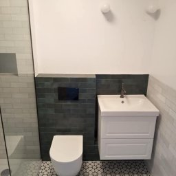 Nowoczesna łazienka po remoncie: biała umywalka z szafką, podwieszana toaleta, ciemnozielone płytki na ścianie i wzorzysta podłoga. Minimalistyczne oświetlenie.