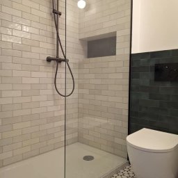 Nowoczesna kabina prysznicowa z ciemną armaturą i geometryczną posadzką. Ściany wyłożone białymi, cegiełkowymi płytkami, akcent ciemnej ściany z toaletą w tle.