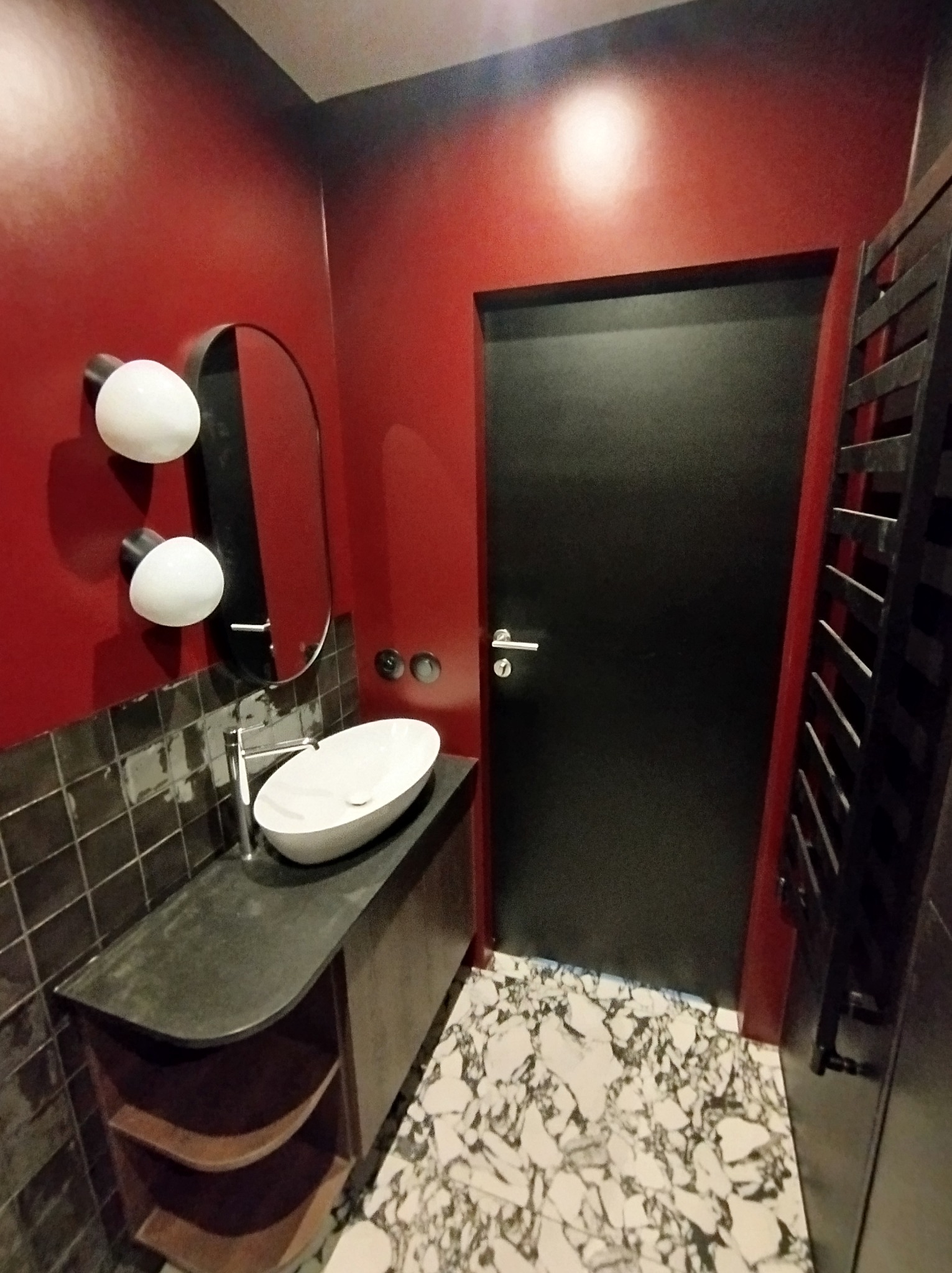 Nowoczesna łazienka z czerwonymi ścianami, czarną armaturą i marmurową podłogą. Umywalka nablatowa, owalne lustro i designerskie oświetlenie tworzą unikalny klimat.