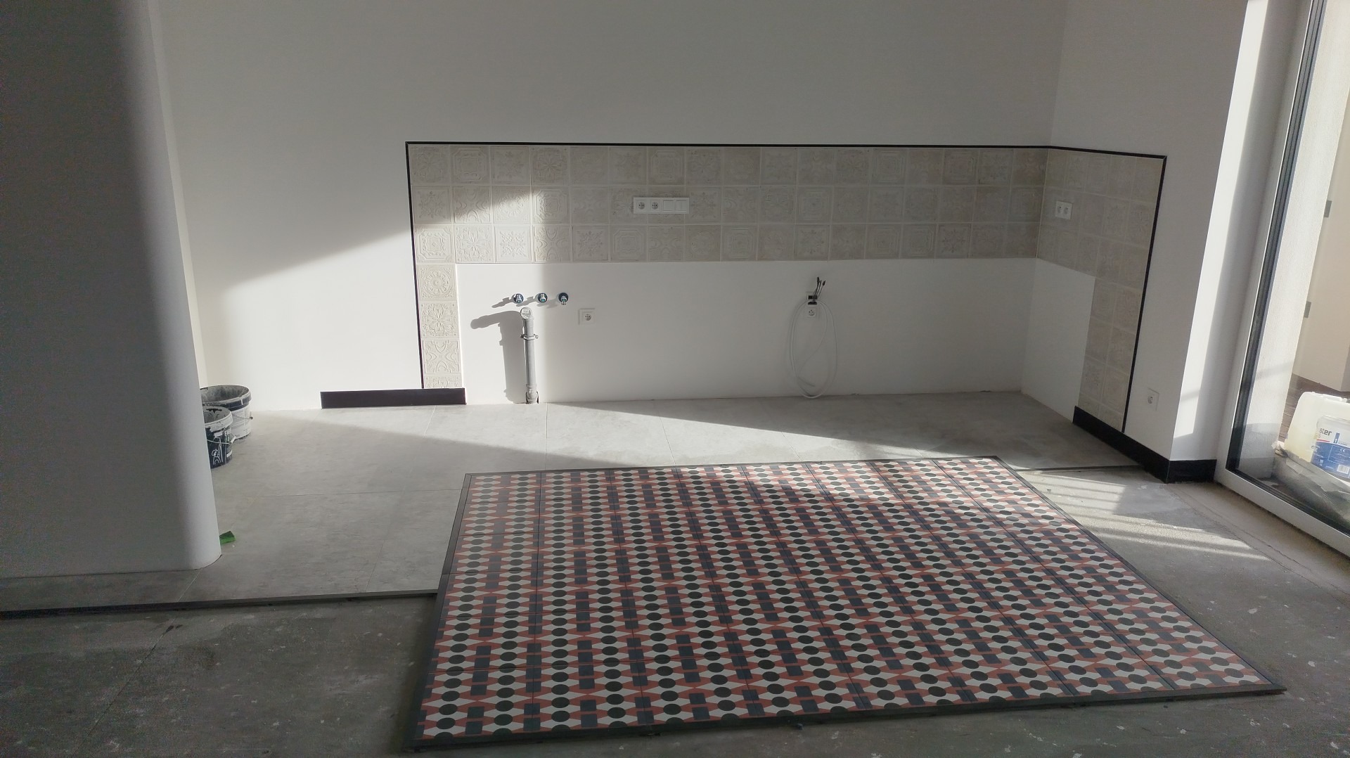 Wnętrze kuchni w trakcie prac wykończeniowych: ułożona podłoga z wzorzystych płytek, częściowo wykończona ściana nad blatem z dekoracyjnymi kaflami i widoczne instalacje.