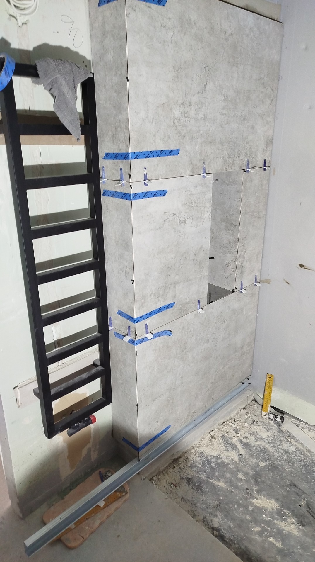 Montaż szarych płytek imitujących kamień na ścianie, z użyciem niebieskiej taśmy i plastikowych krzyżyków. Widoczna drabina i profil podłogowy.