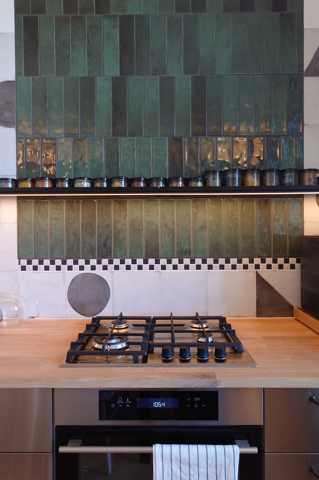 Nowoczesna kuchnia z zielonymi, pionowymi płytkami nad blatem i rzędem słoiczków z przyprawami. Nietypowe wzory płytek i drewniany blat dodają unikalnego charakteru.