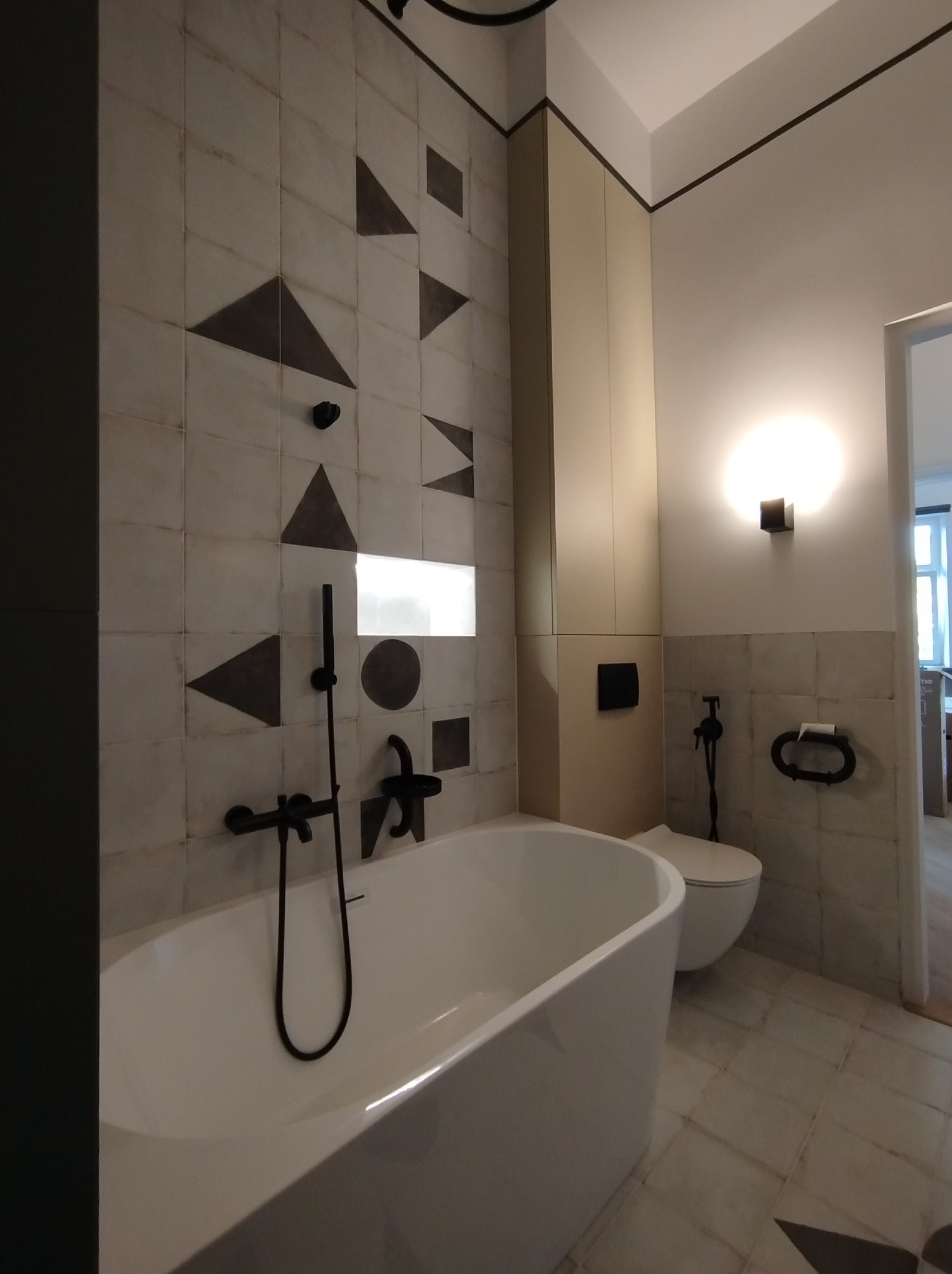 Nowoczesna łazienka z białą wanną, czarną armaturą i geometrycznymi wzorami na płytkach. Uwagę przykuwa minimalistyczny design i stonowana kolorystyka.