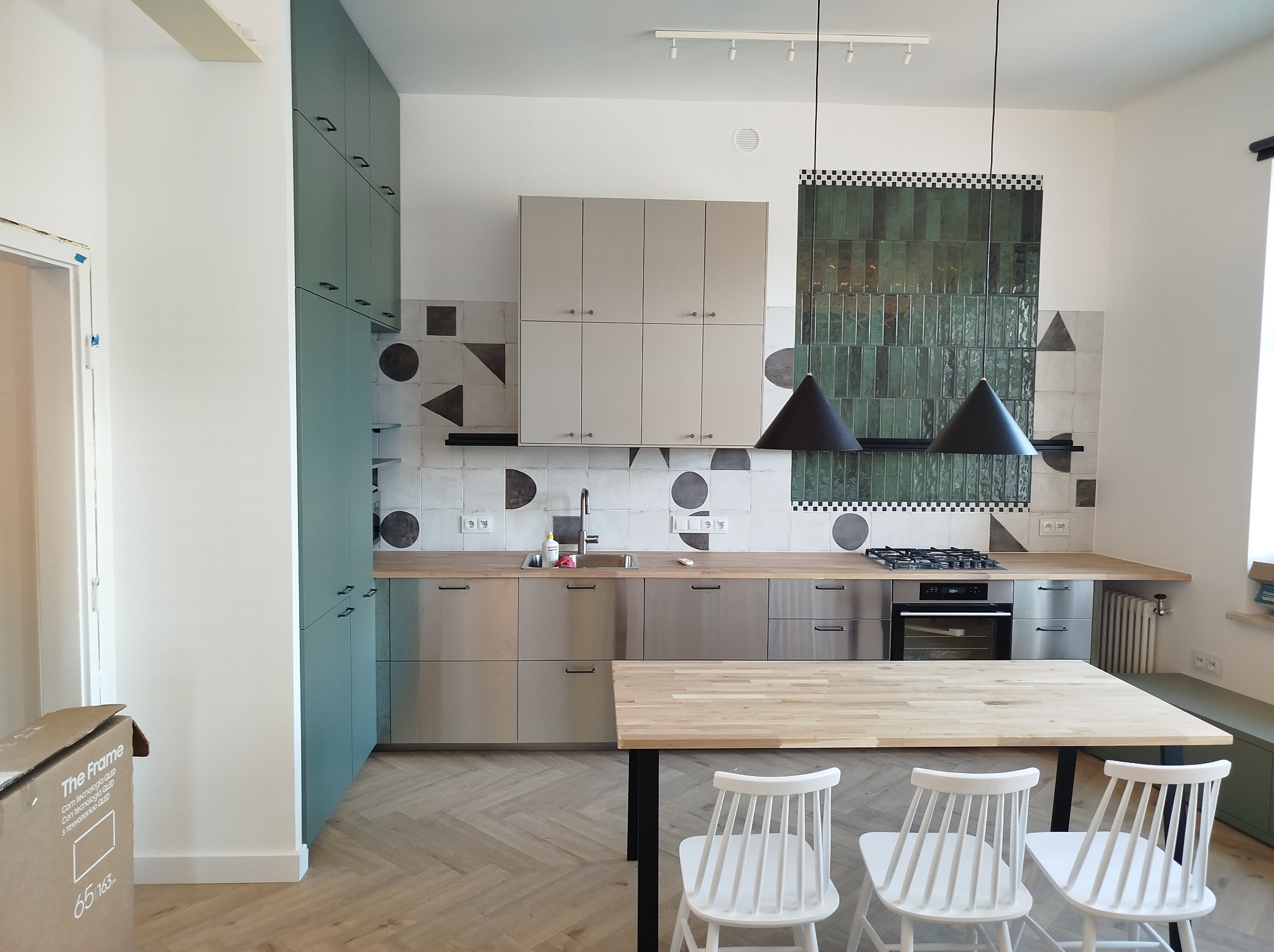 Nowoczesna kuchnia z zielonymi i beżowymi szafkami, drewnianym blatem i stołem. Na ścianie dekoracyjne płytki w geometryczne wzory i zielona mozaika.