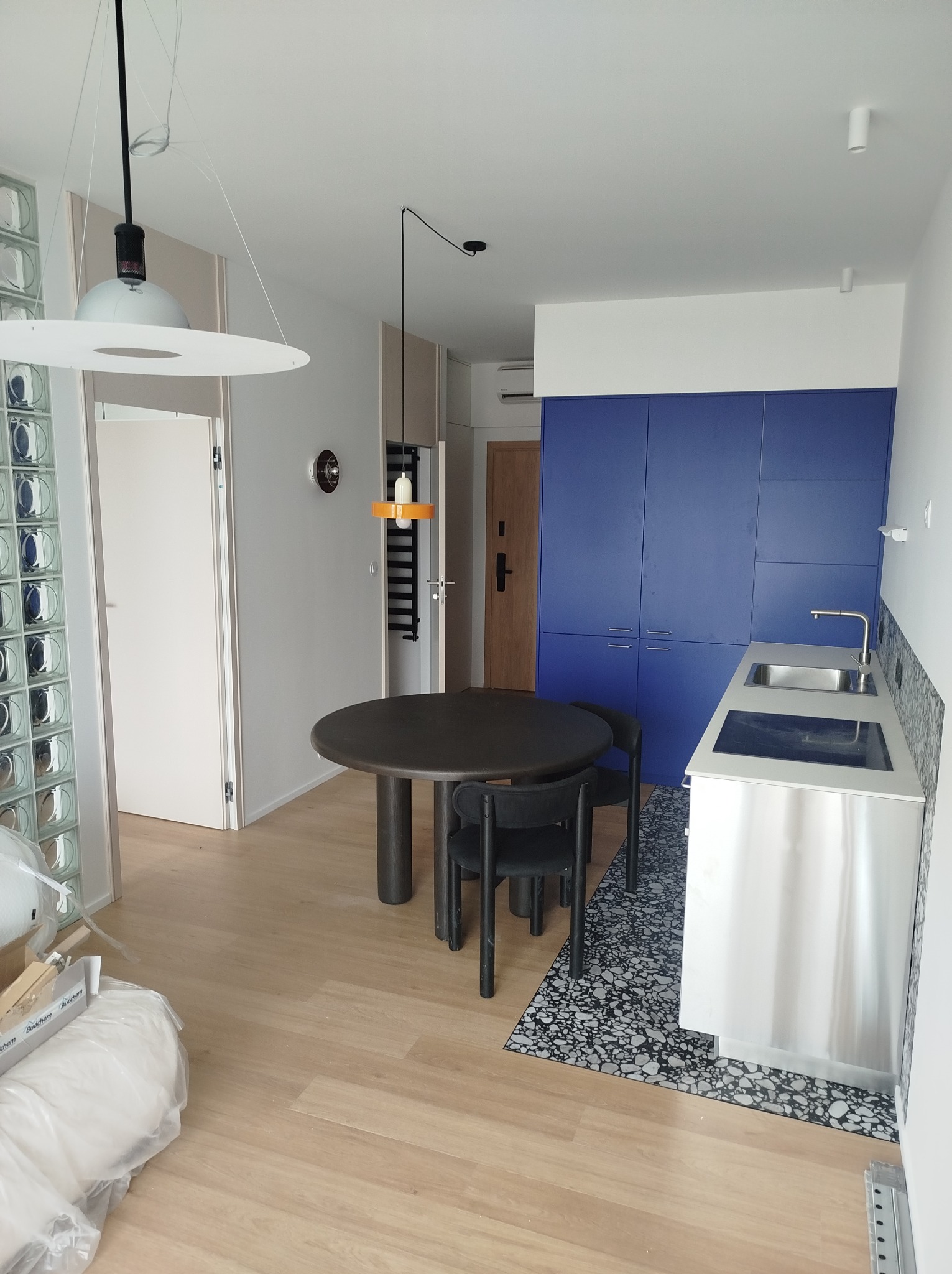 Nowoczesne wnętrze mieszkania: okrągły stół z krzesłami, zabudowa kuchenna w kolorze niebieskim, parkiet i dekoracyjna ściana z luksferów. Widoczne lampy wiszące.