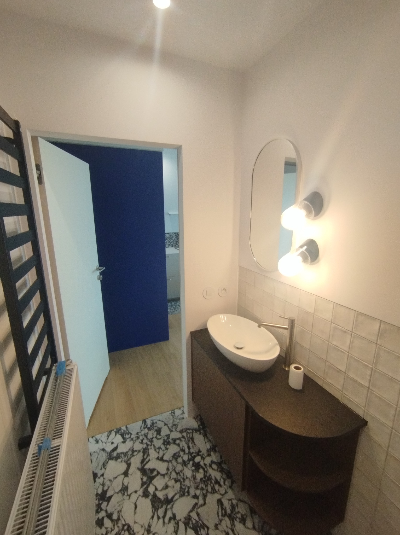 Nowoczesna łazienka z owalną umywalką nablatową, lustrem z oświetleniem i grzejnikiem drabinkowym na tle białych ścian i wzorzystej podłogi.