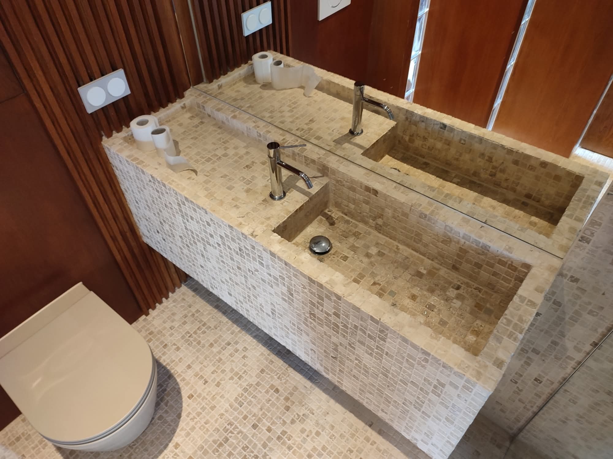 Nowoczesna łazienka z beżową mozaiką: umywalka nablatowa z dwoma kranami, lustro, toaleta. Drewniane panele na ścianie dodają ciepła. Widok z góry.