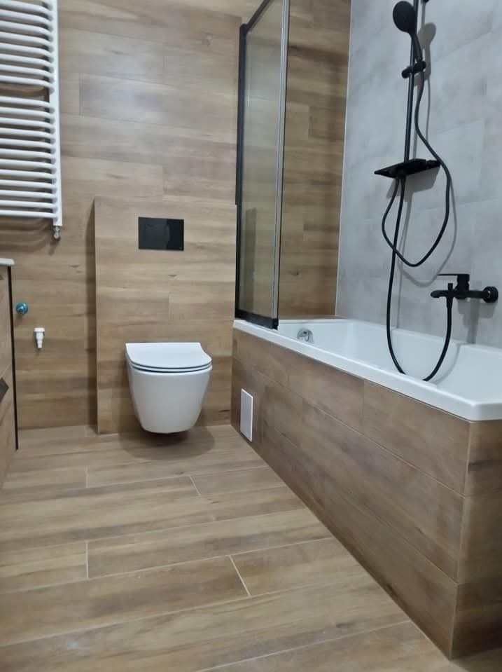 Nowoczesna łazienka z drewnopodobnymi płytkami, białą armaturą i czarną baterią prysznicową. Kabina prysznicowa z czarnymi profilami. Minimalistyczny design.