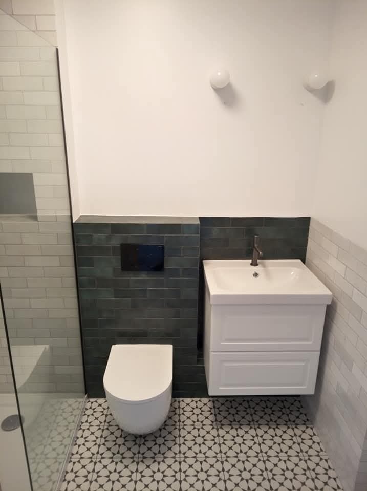 Nowoczesna łazienka po remoncie: biała umywalka z szafką, podwieszana toaleta, ciemnozielone płytki na ścianie i wzorzysta podłoga. Minimalistyczne oświetlenie.