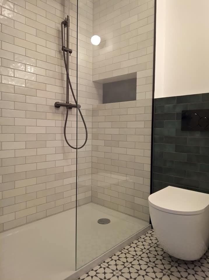 Nowoczesna kabina prysznicowa z ciemną armaturą i geometryczną posadzką. Ściany wyłożone białymi, cegiełkowymi płytkami, akcent ciemnej ściany z toaletą w tle.
